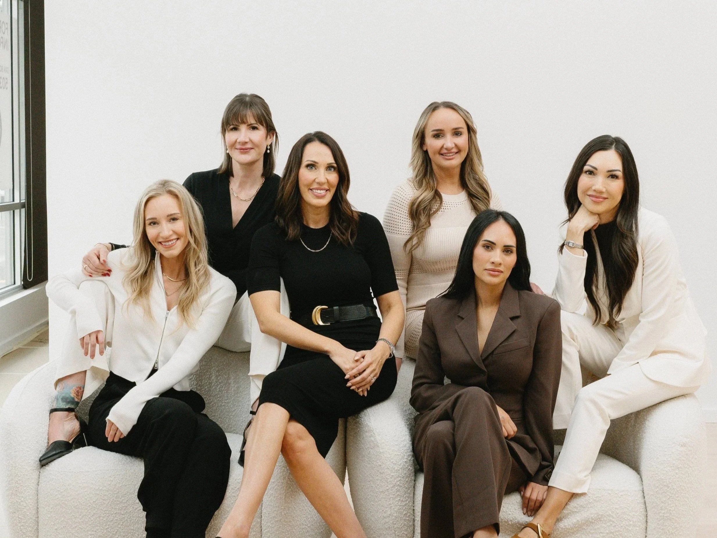 Seven women sitting and standing on a white couch in front of a plain white wall, smiling, dressed in business casual attire.