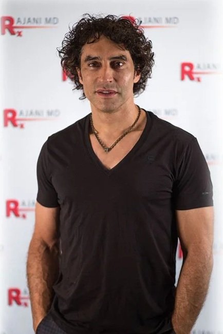 Man with curly hair wearing a black v-neck t-shirt and a beaded necklace standing in front of a white background with red and black text.