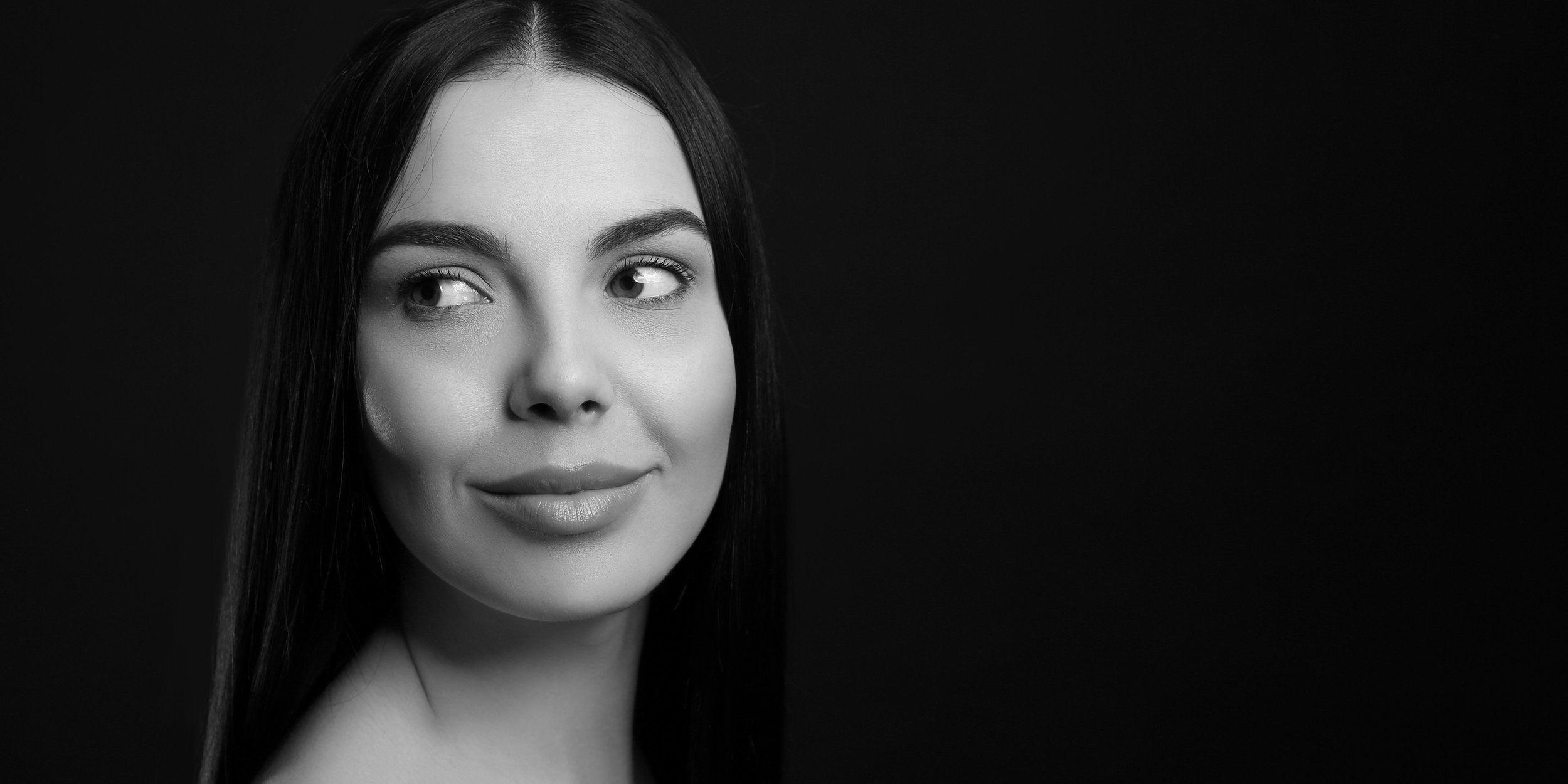Black and white portrait of a woman with long dark hair, smiling softly, looking slightly to her left, against a dark background.