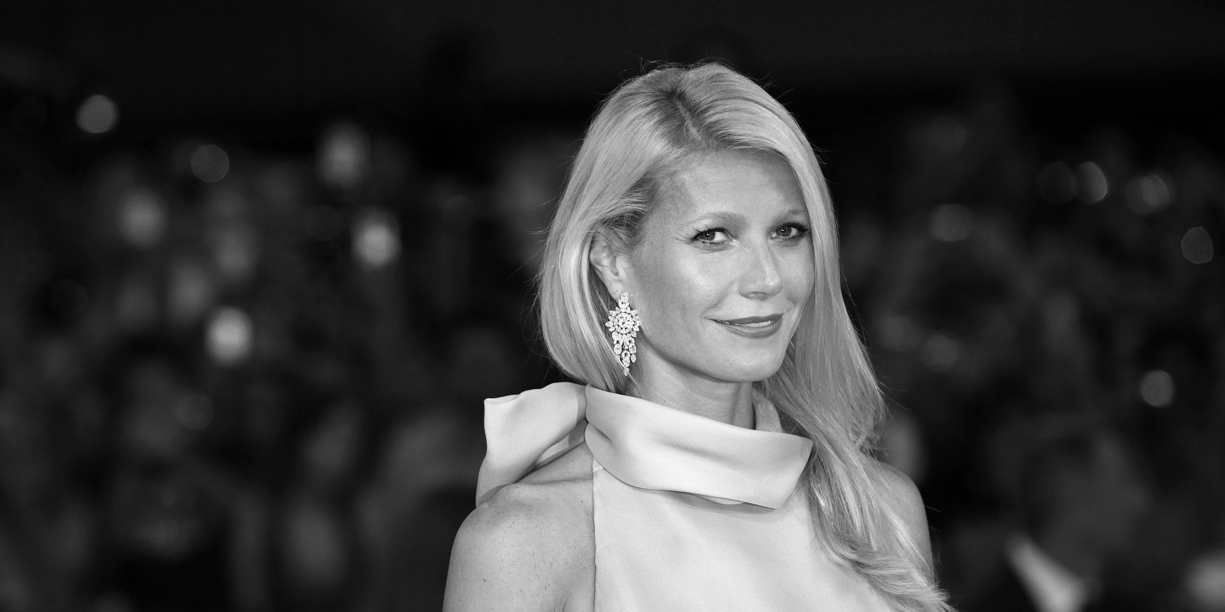 Black and white portrait of a woman with long blonde hair, wearing elegant earrings and a high-neck dress, at night with blurred lights in the background.