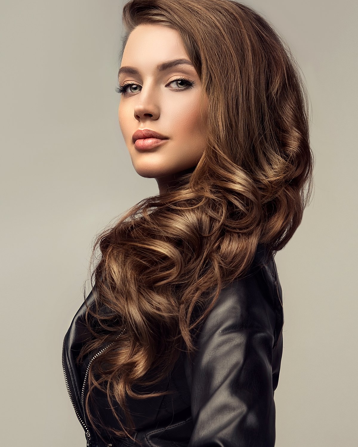 A woman with long, wavy, brown hair looking over her shoulder, wearing a black leather jacket.