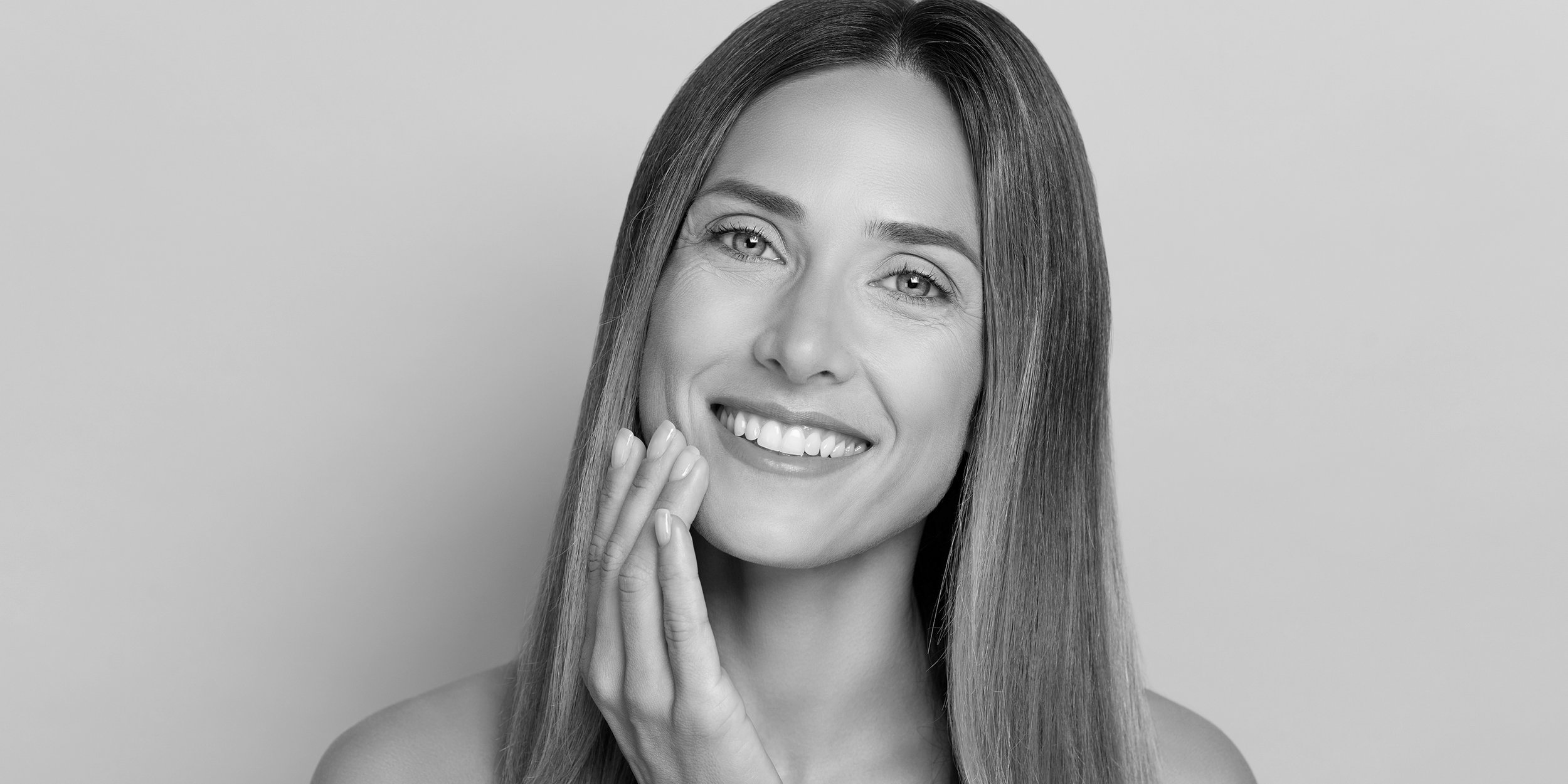 Black and white portrait of a smiling woman with long straight hair touching her cheek, on a plain background.