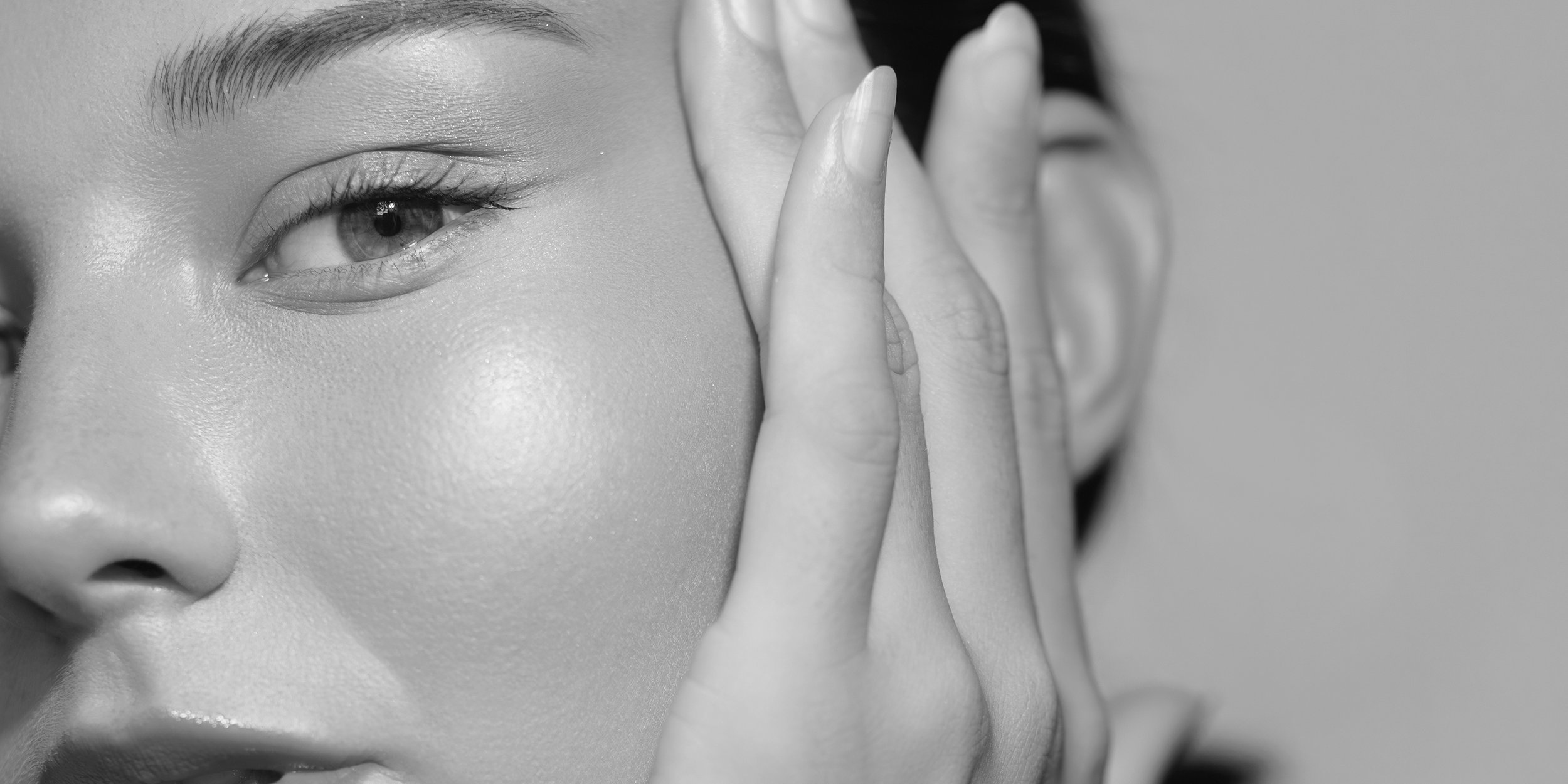Black and white close-up of a person's face, focusing on one eye and cheek, with hand covering part of their face and fingers touching their cheek.