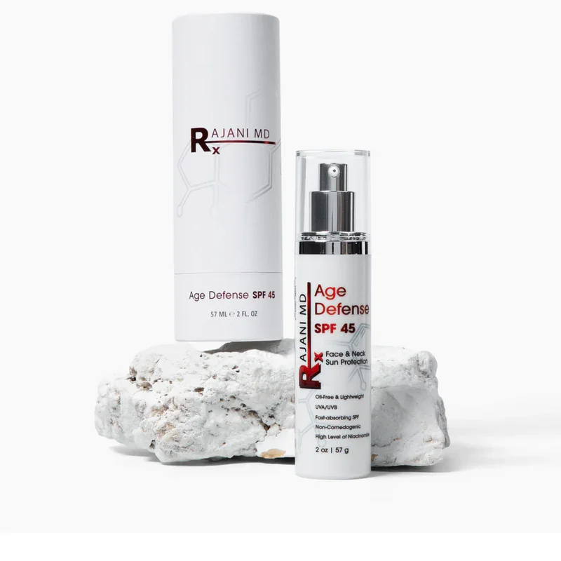 A white bottle and a white tube of Rajani MD Age Defense SPF 45 sunscreen are displayed on a piece of white coral against a white background.
