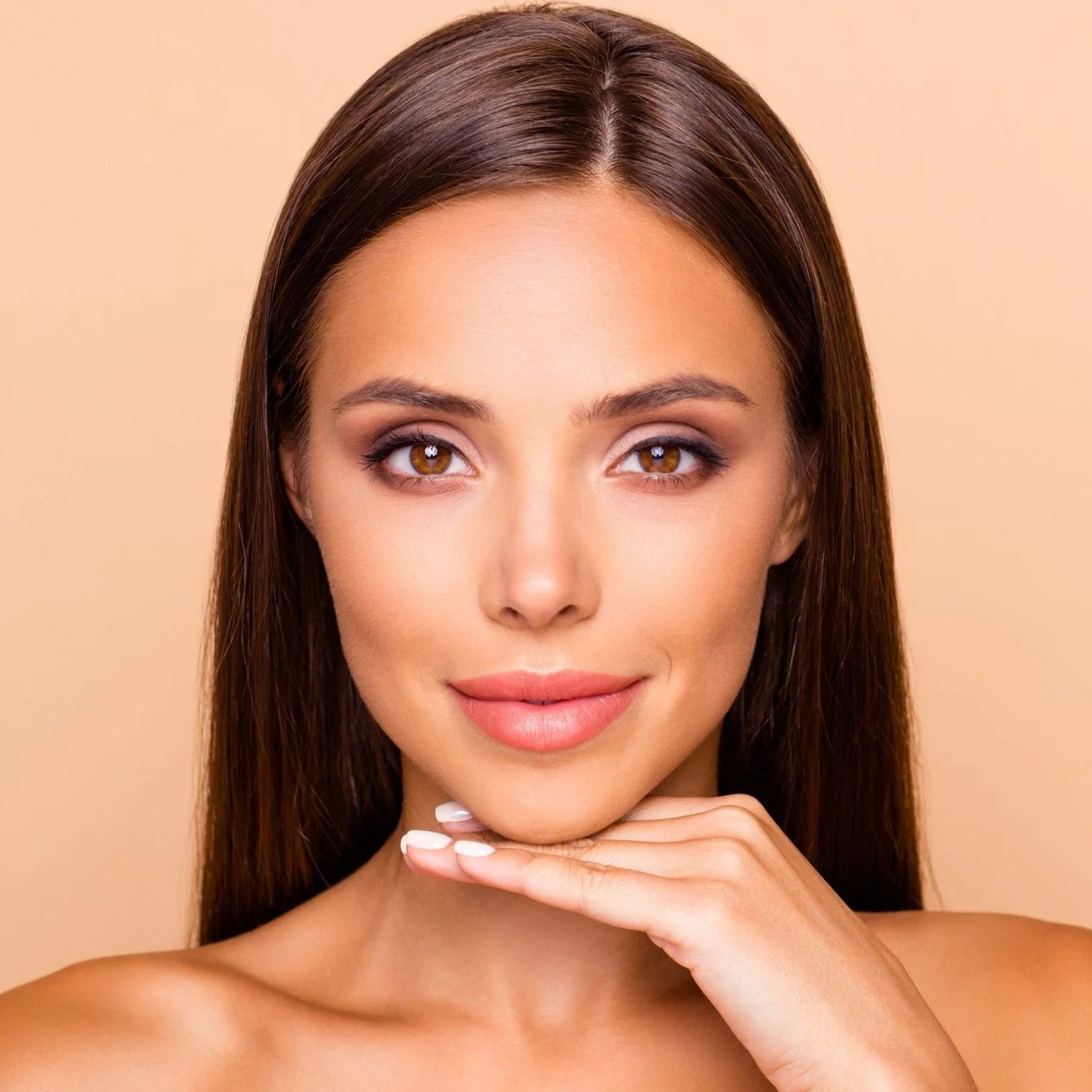 Close-up of a woman with brown hair and makeup, resting her chin on her hand against a beige background.