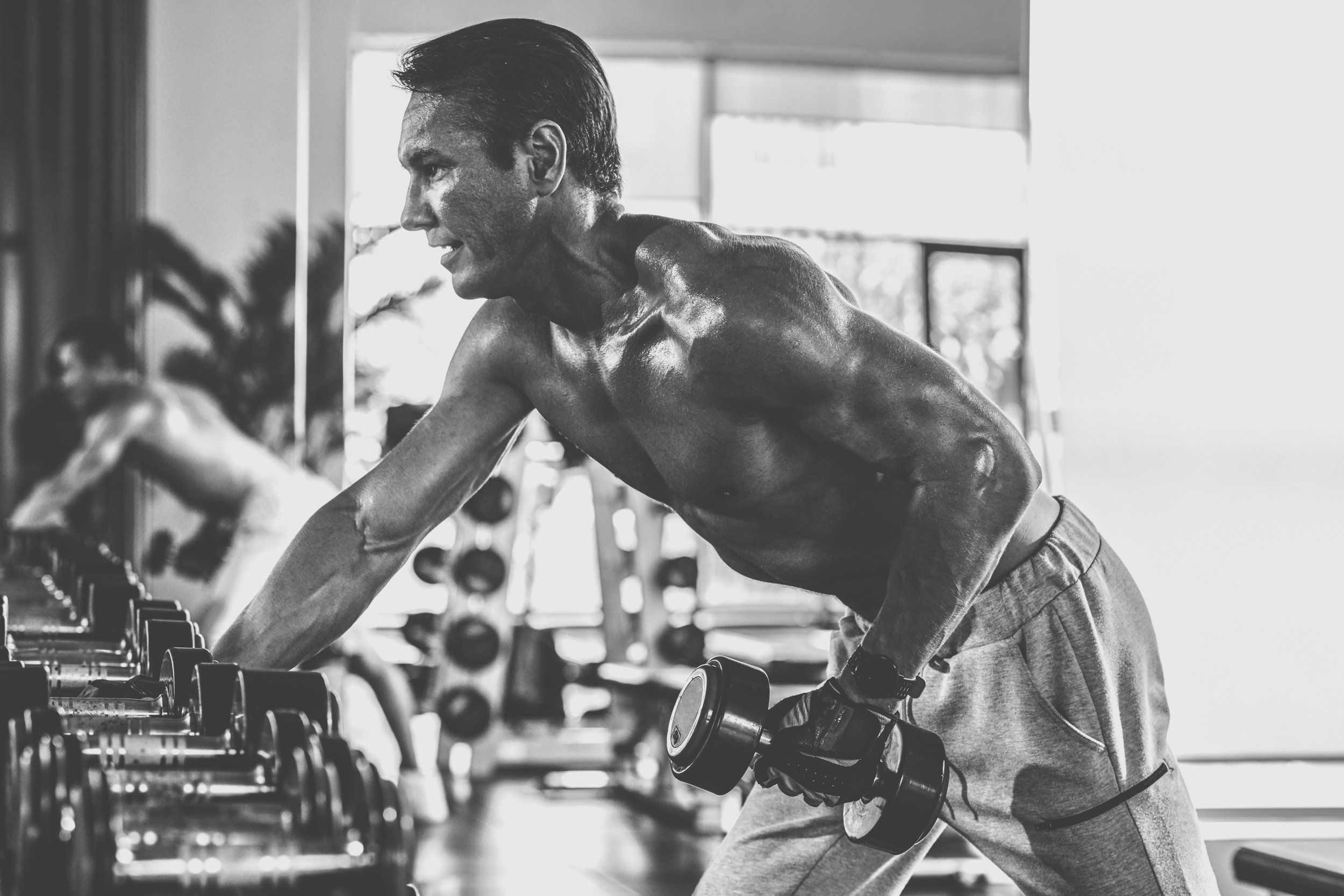 A shirtless man working out with dumbbells at the gym, focused, in black and white, with workout equipment and another person in the background.