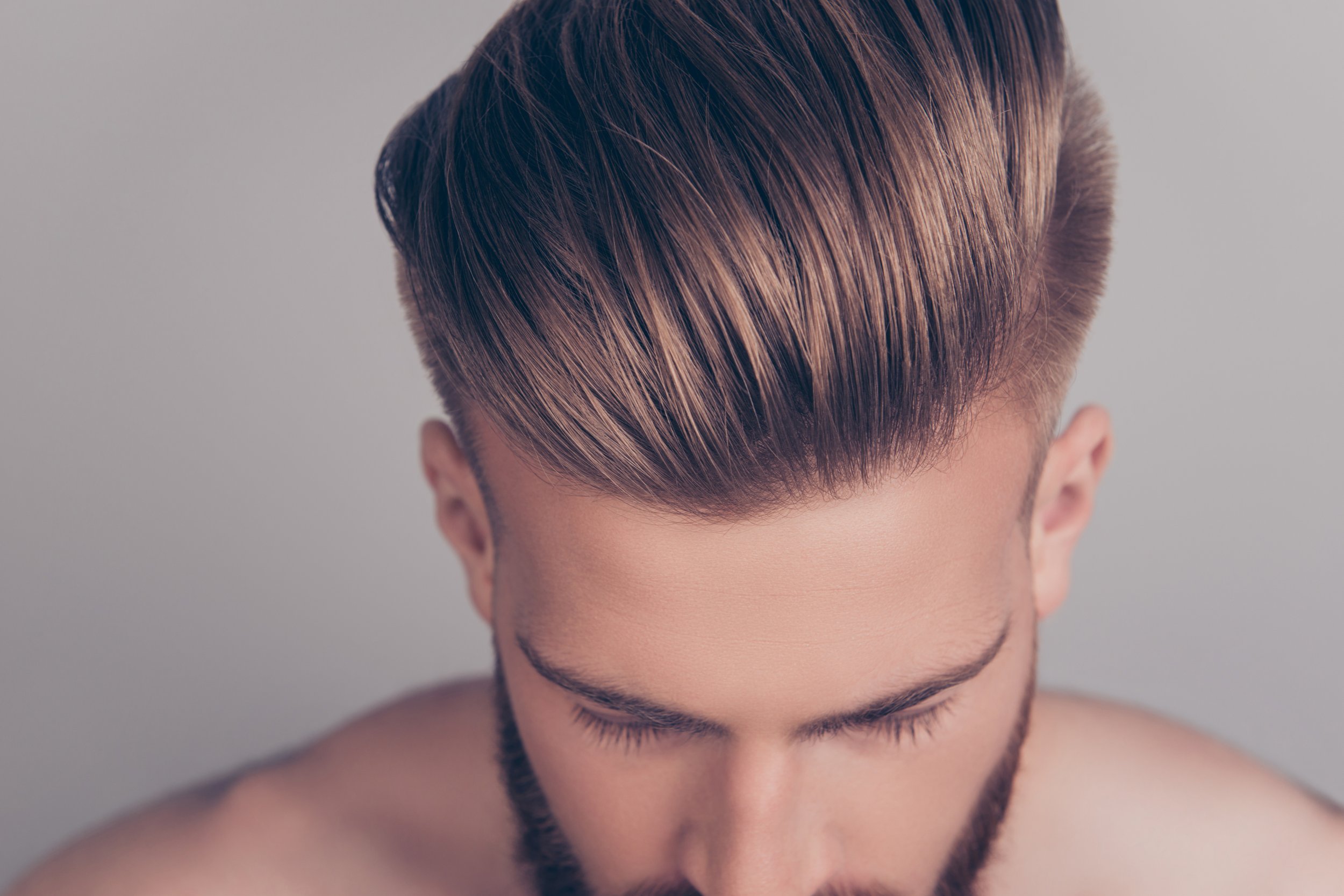 Close-up of a young man with styled, light brown hair and a beard, looking downward, with a neutral background.