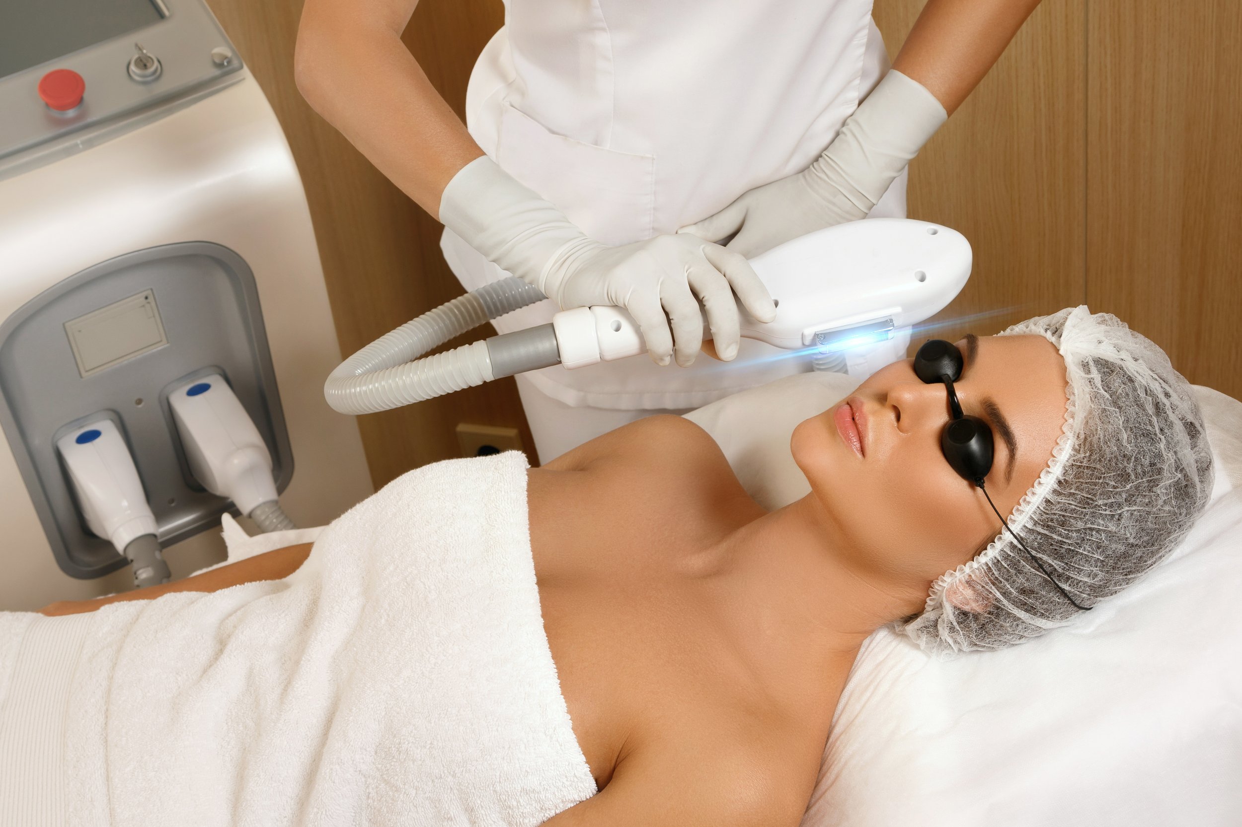 A woman receiving a laser skin treatment while lying on a bed, wearing protective goggles and a hair cap, with a technician holding a laser device.