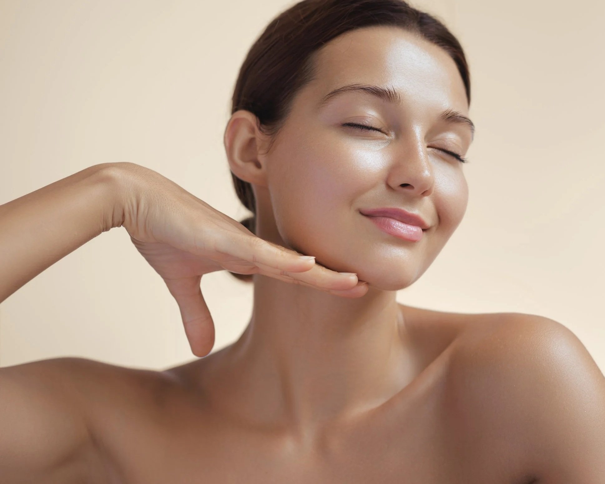 Close-up of a smiling woman with clear, glowing skin, resting her chin on her hand, with bare shoulders against a neutral background.