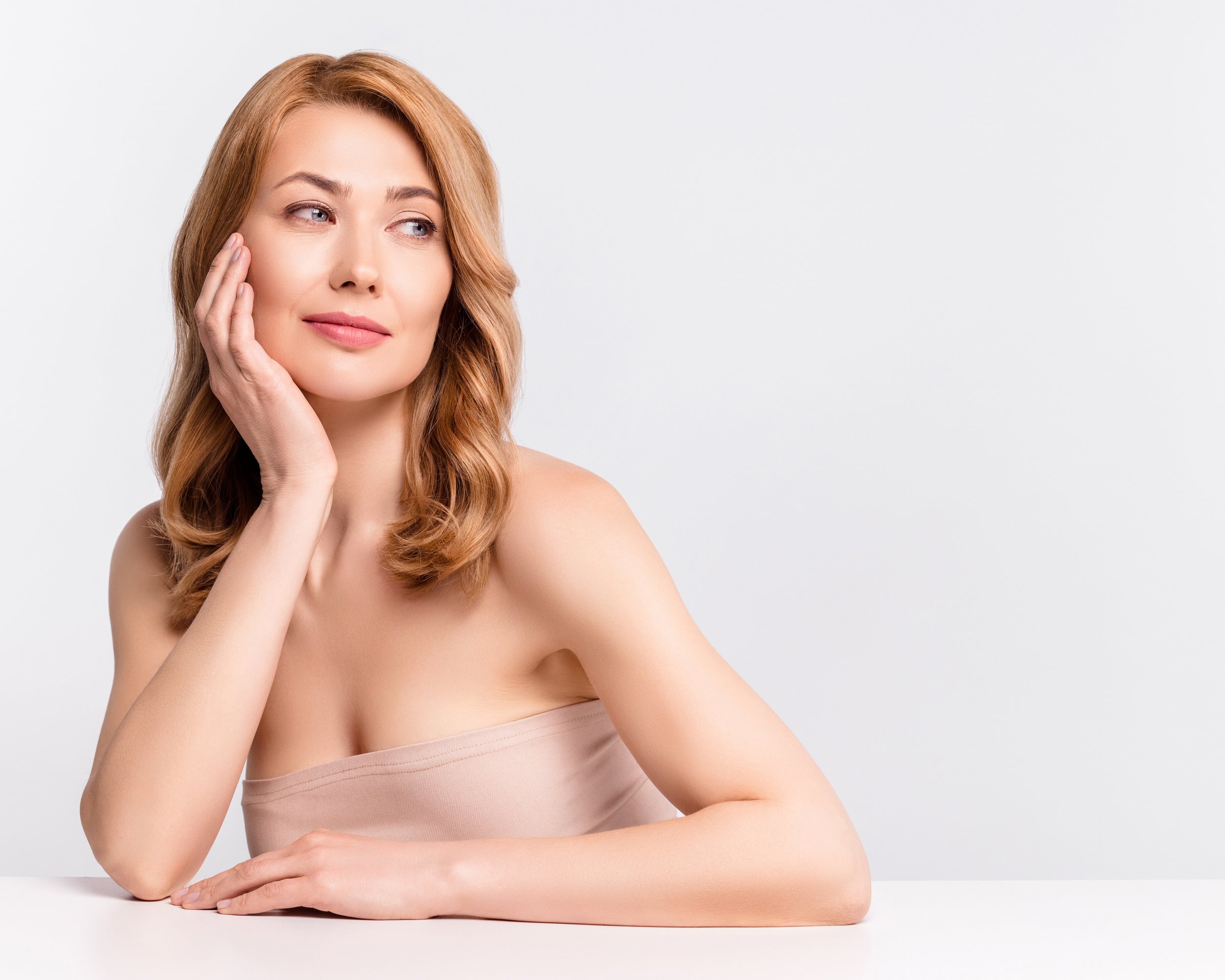 A woman with shoulder-length I-wavy red hair, resting her head on her left hand, looking thoughtfully to her right, sitting against a plain white background.