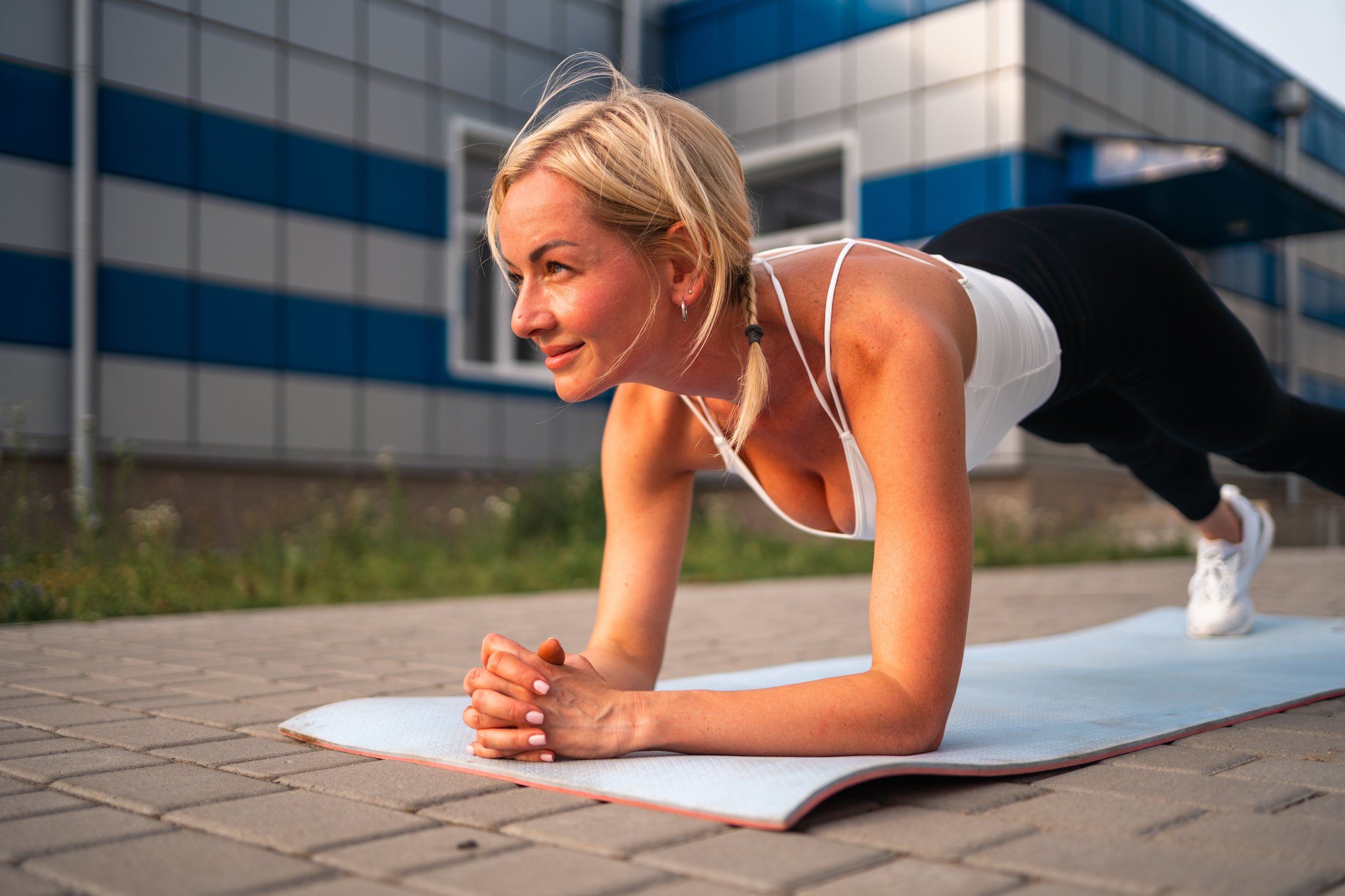 Woman doing plank exercise outdoors on a yoga mat in front of a modern building.