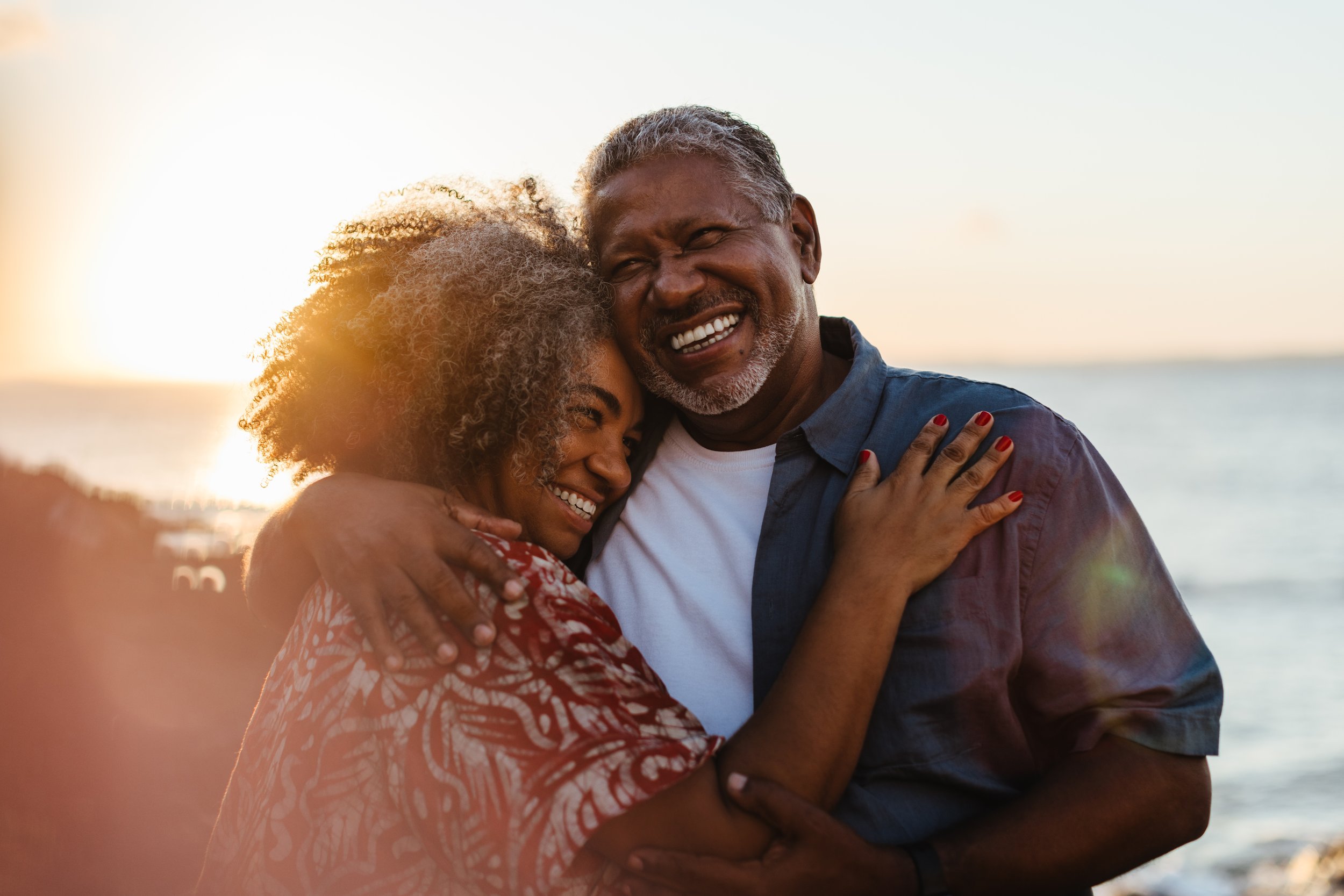 A joyful older couple hugging each other on a beach at sunset, smiling happily.
